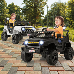 Two children play with black and white ride-on toyJeep Wrangler-style vehicles outdoors on a paved park path, with trees and greenery in the background.