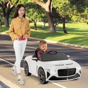 A woman walks beside a young boy driving a white convertible ride-on toy car on a paved path in a park. The toy car is styled like a sports car with black wheels, a black windshield, and a front grille.