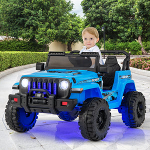 A young child sitting in a bright blue ride-on toy jeep designed to look like a Jeep Wrangler, parked outdoors on a paved driveway with green trees and hedges in the background.