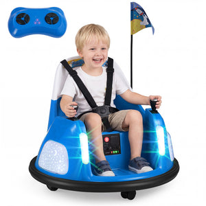 A young boy wearing a white t-shirt and khaki shorts is smiling while riding a blue and black toy race car with integrated seatbelts and two joysticks. A blue remote control is shown in the top left corner, and a small flag is attached to the roof of the car.