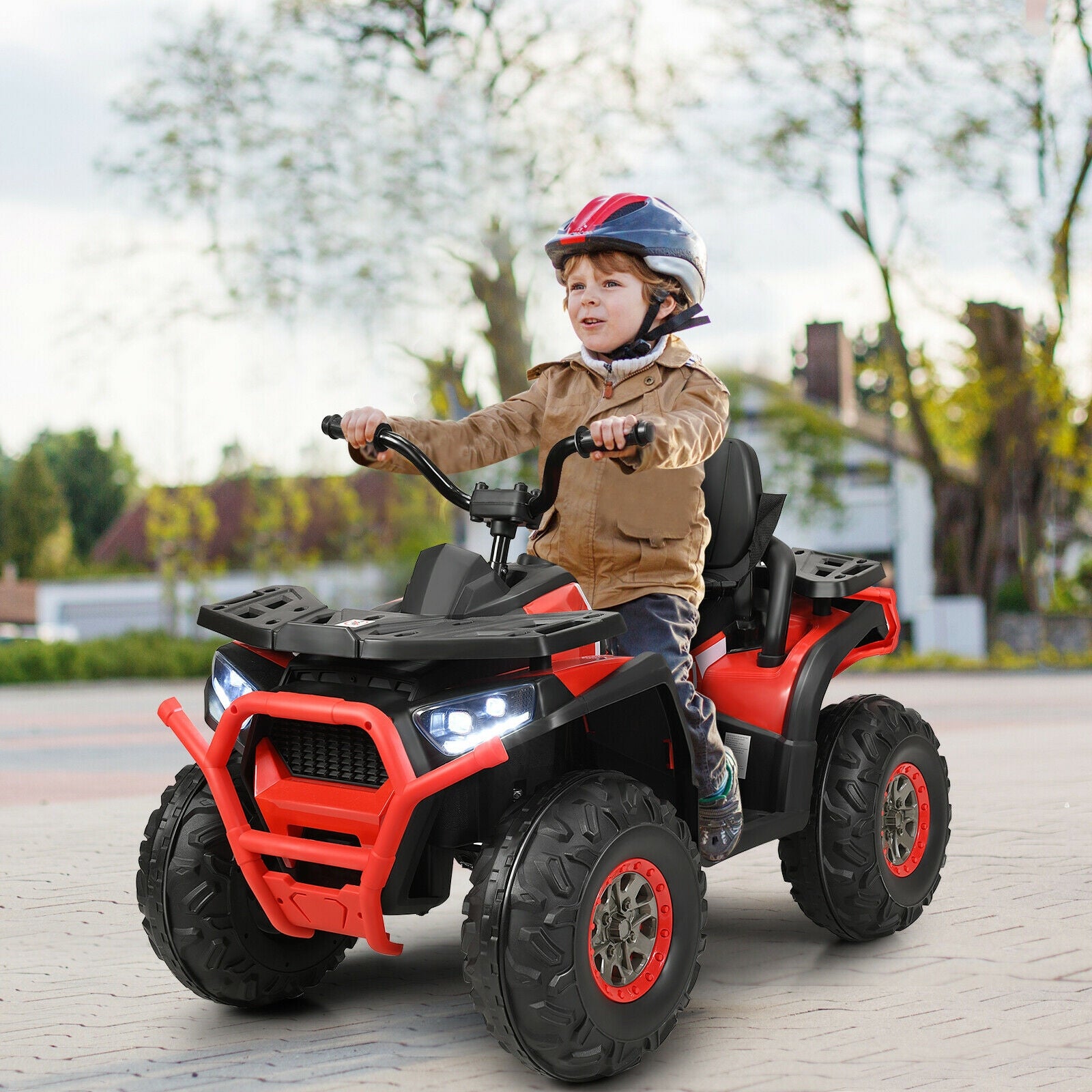 Child riding a red and black toy ATV on a paved path with trees in the background