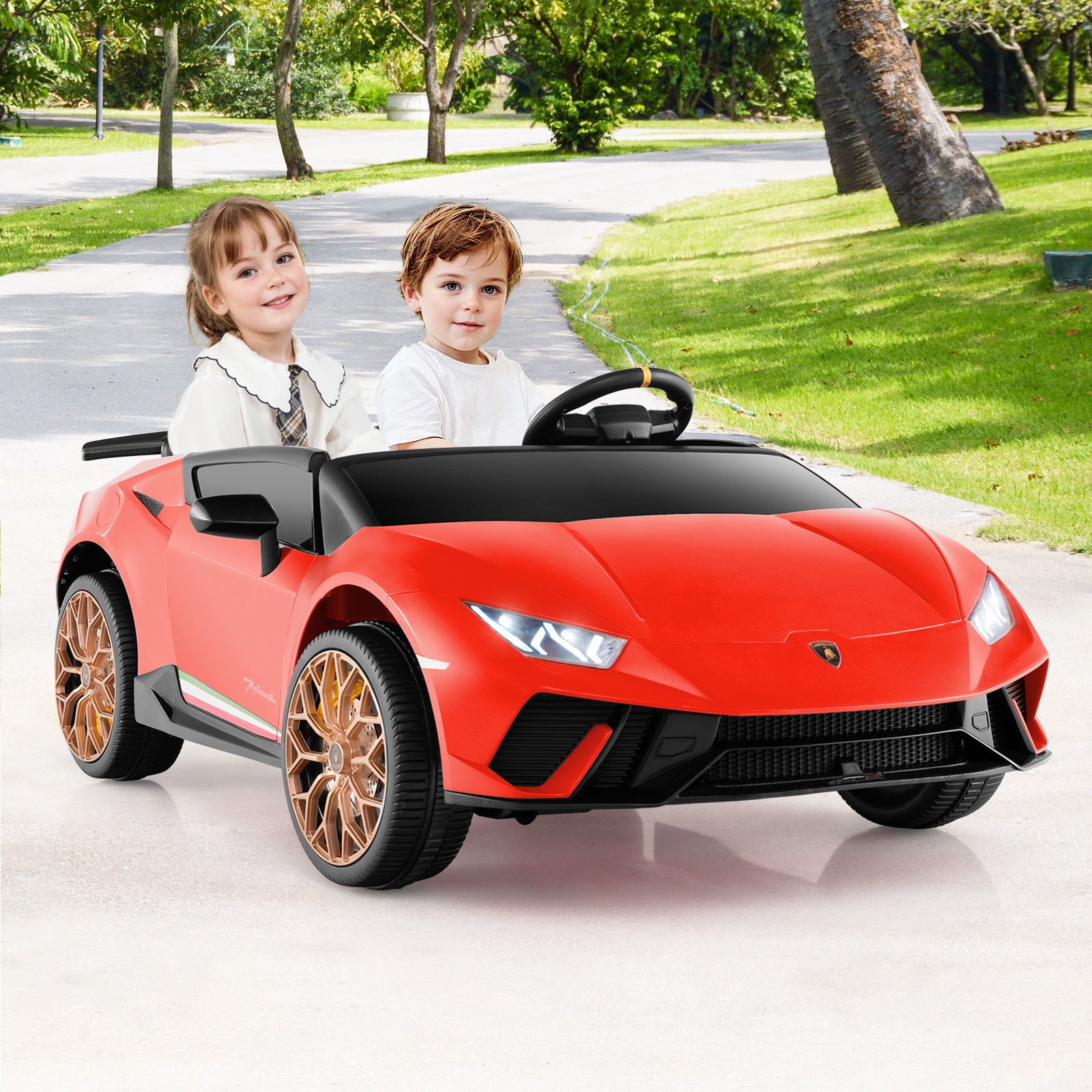 Two children sitting in a red toy car on a driveway with greenery in the background
