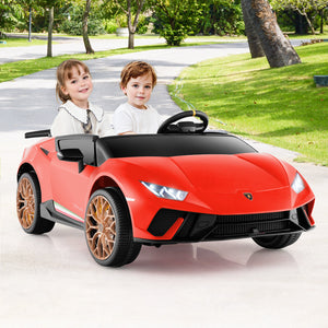 Two children sitting in a red toy car on a driveway with greenery in the background