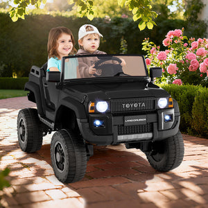 Two children in a toy Toyota vehicle on a brick path with flowers and greenery in the background