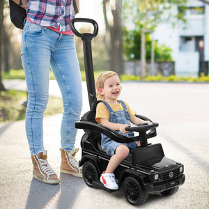 Child in a toy car being pushed by an adult on a path with trees and buildings in the background