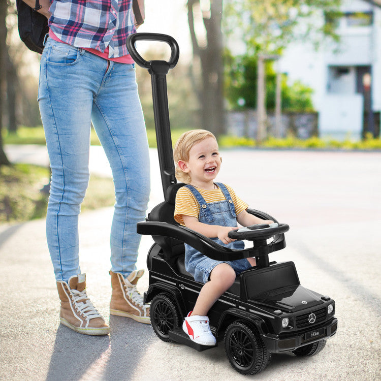 Child in a toy car being pushed by an adult on a path with trees and buildings in the background