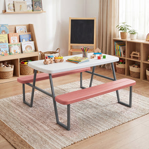 Children's play area with a white table and pink benches in a room with bookshelves and toys.