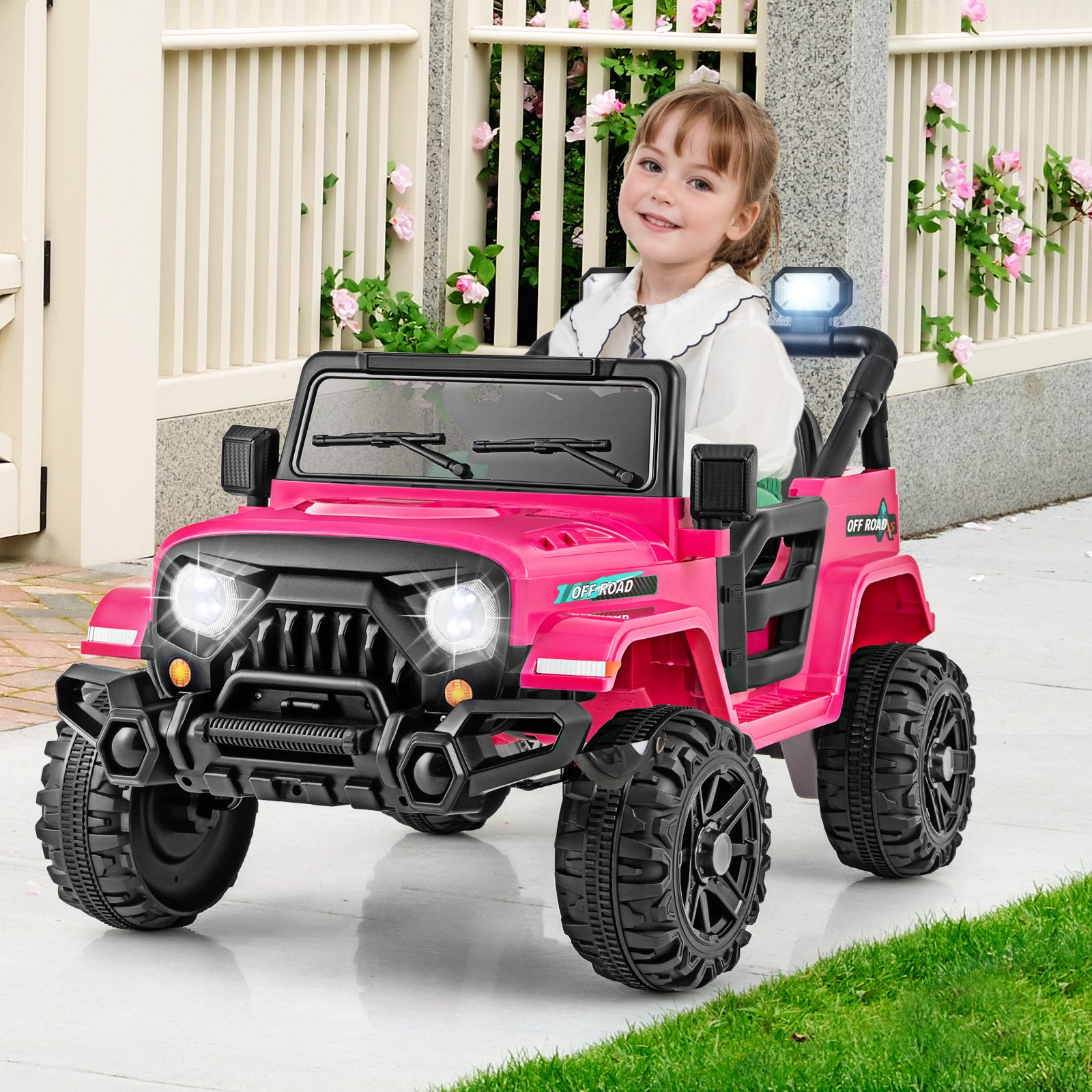 Child sitting in a pink toy jeep with a white fence and flowers in the background