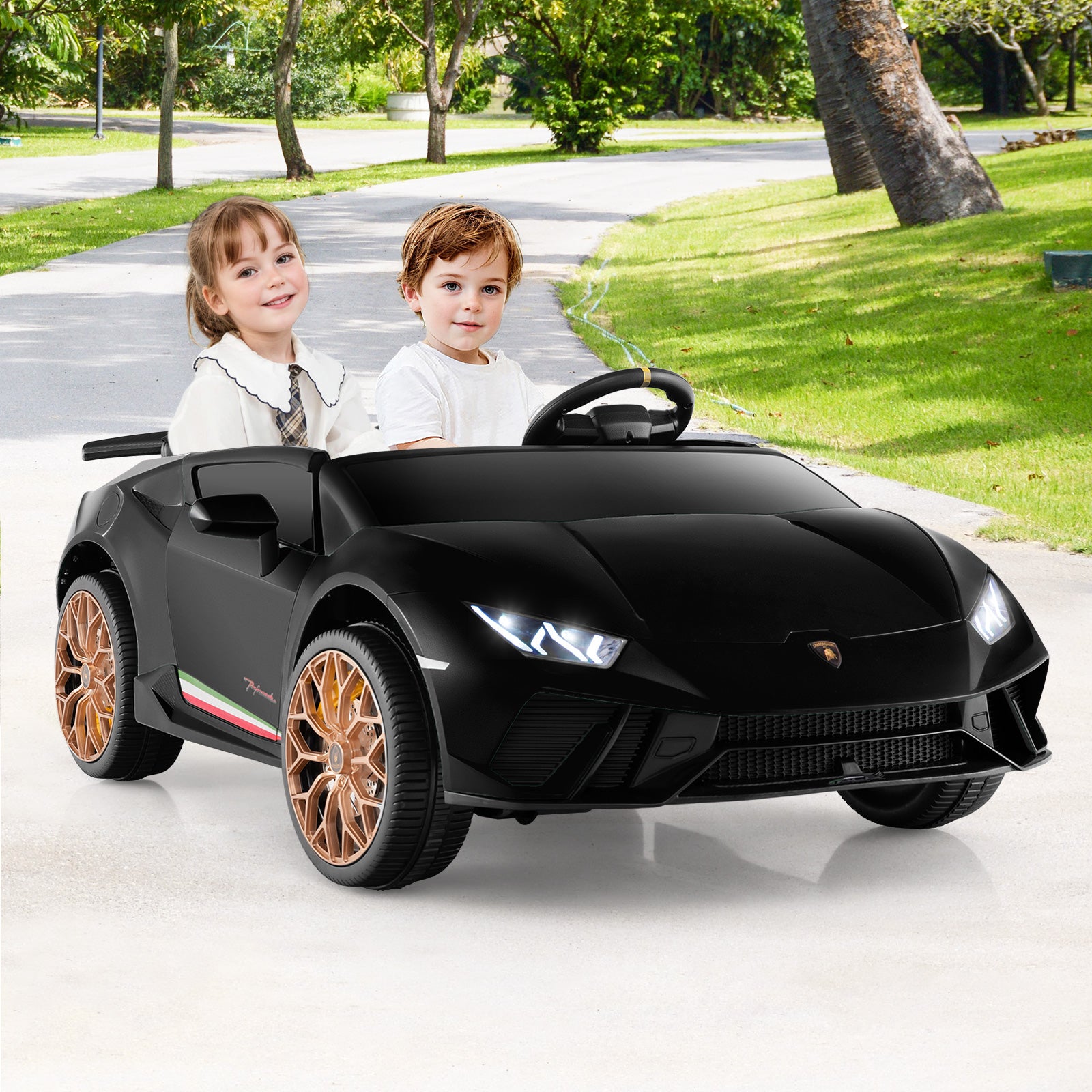 Two children sitting in a black toy car on a driveway with greenery in the background