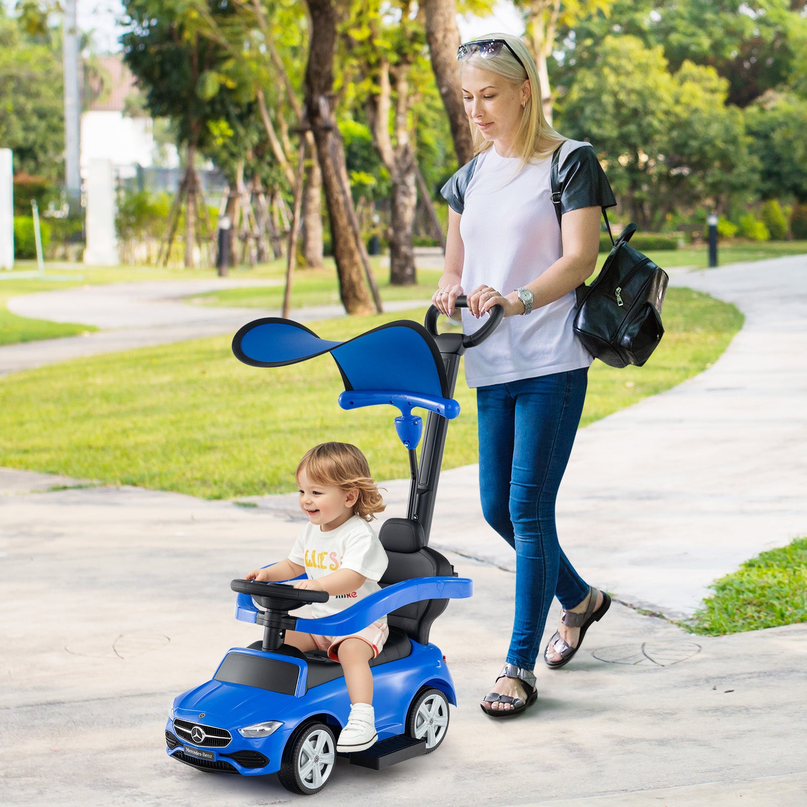 Woman pushing a child in a blue toy car with a steering wheel on a sidewalk.