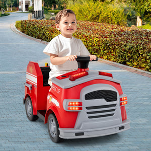 A young child wearing a white t-shirt drives a red and gray toy fire truck on a paved pathway with green hedges in the background.