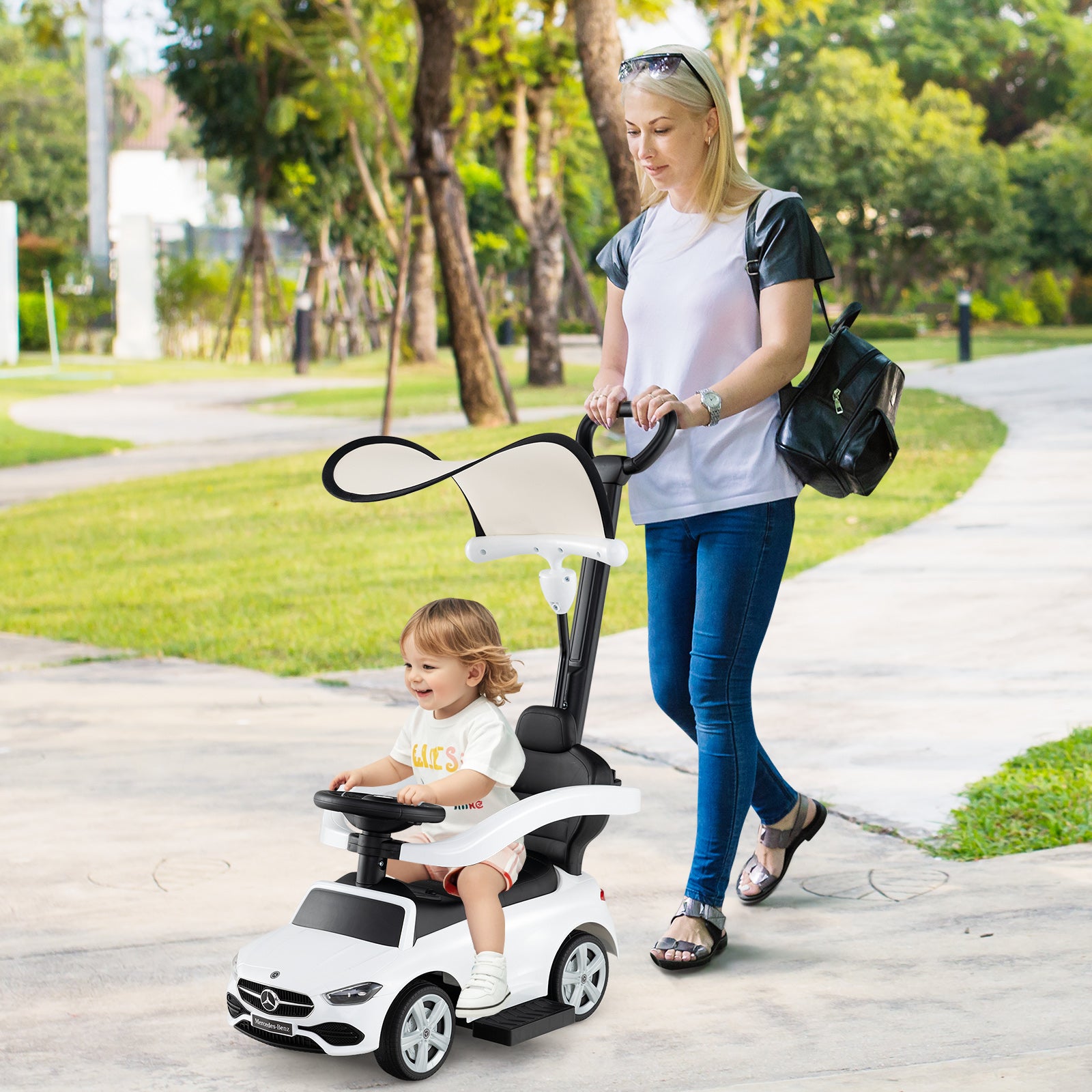 Woman pushing a child in a toy car on a sidewalk with trees in the background