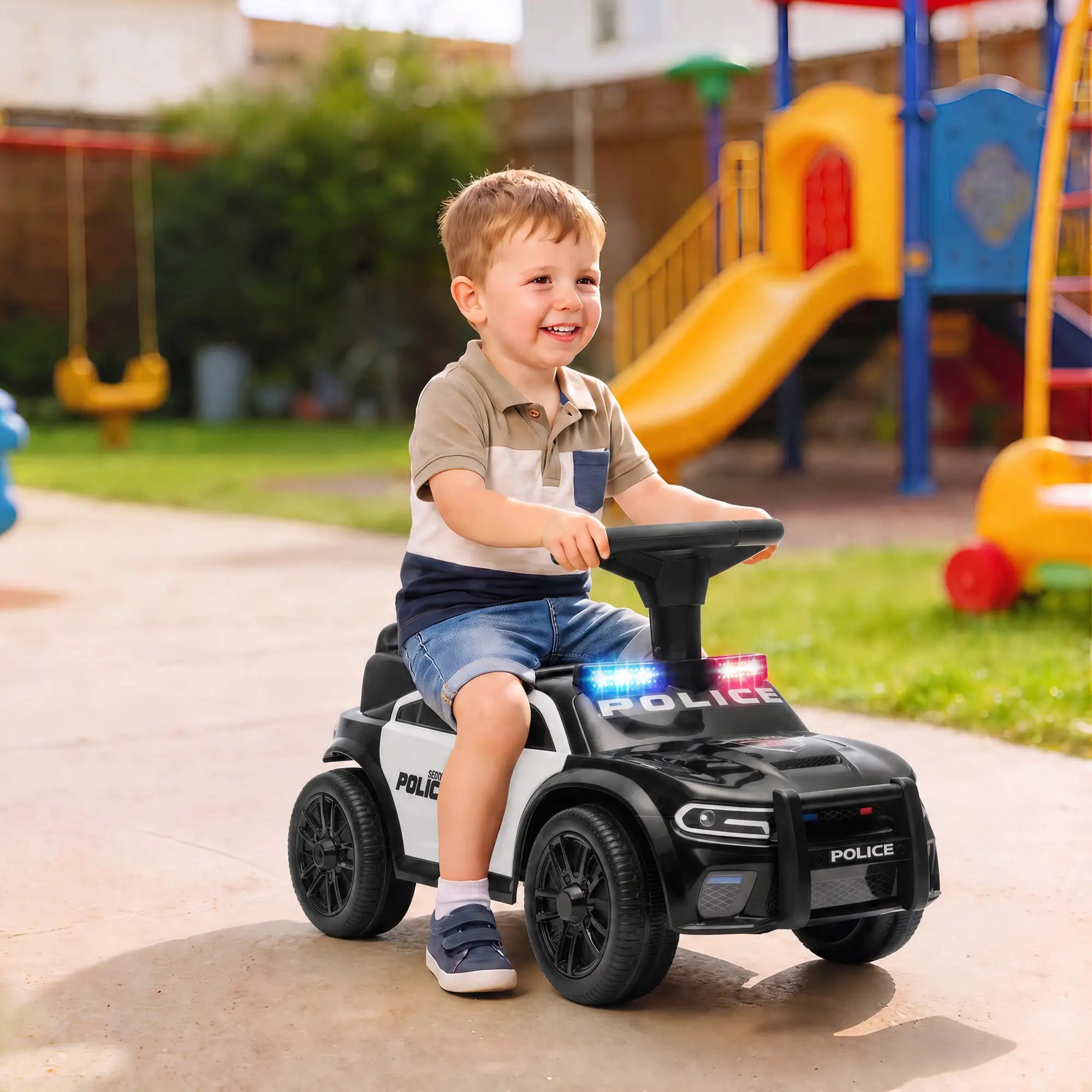 A young boy wearing a beige and navy polo shirt and denim shorts is smiling while riding a black and white police toy car outdoors on a paved playground path.