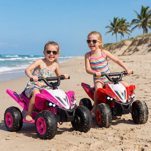 Two young girls riding toy atvs on a sandy beach with palm trees in the background.