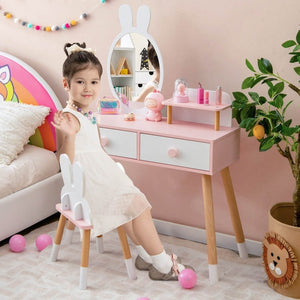 Child sitting on a small stool in front of a pink children's vanity table with mirror and toys.