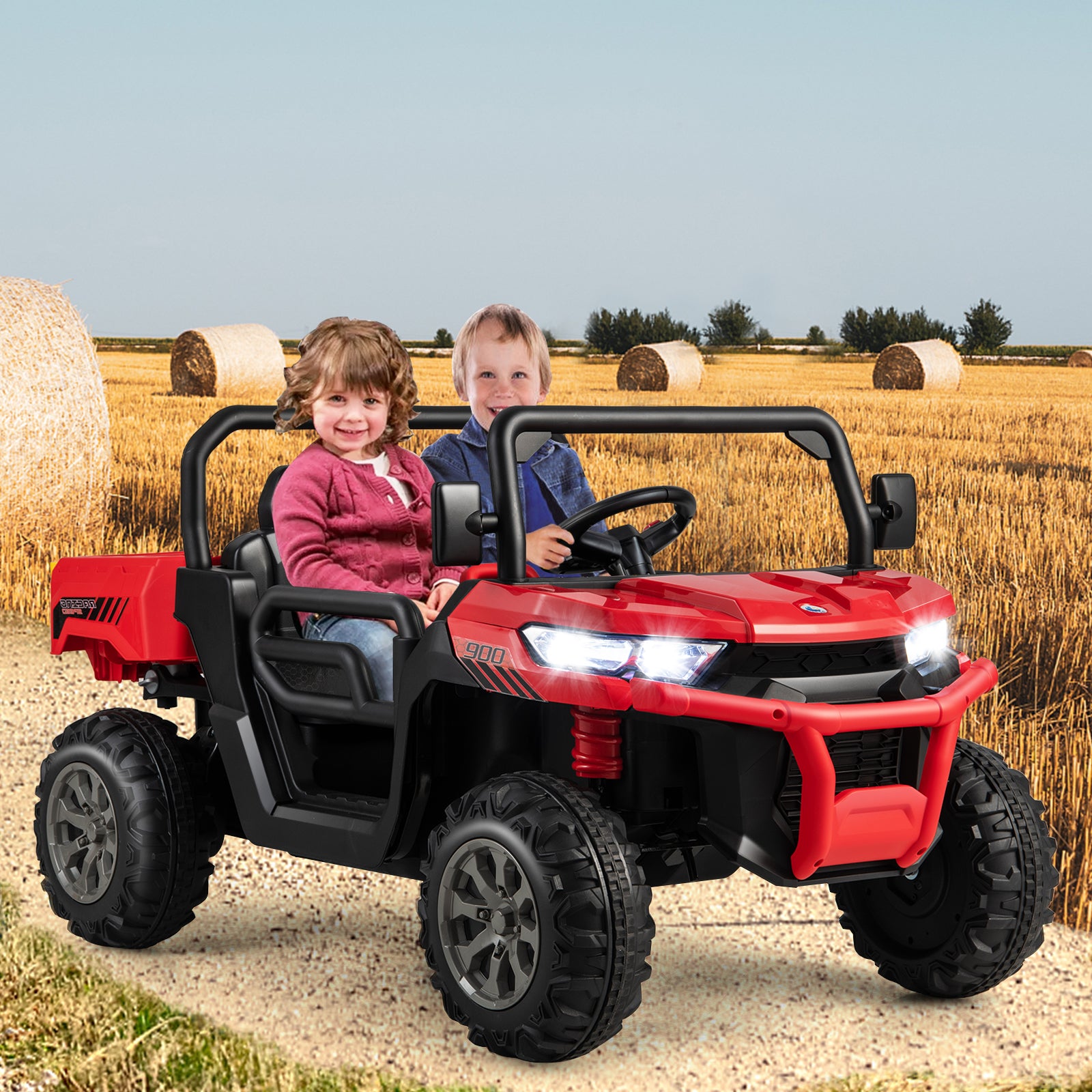 Two children sitting in a red toy off-road vehicle in a field with hay bales.