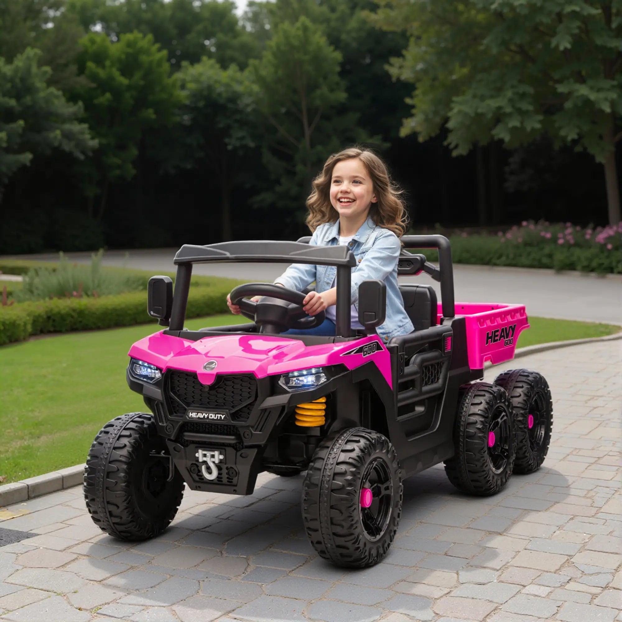 Child sitting in a pink and black toy truck on a paved path with greenery in the background
