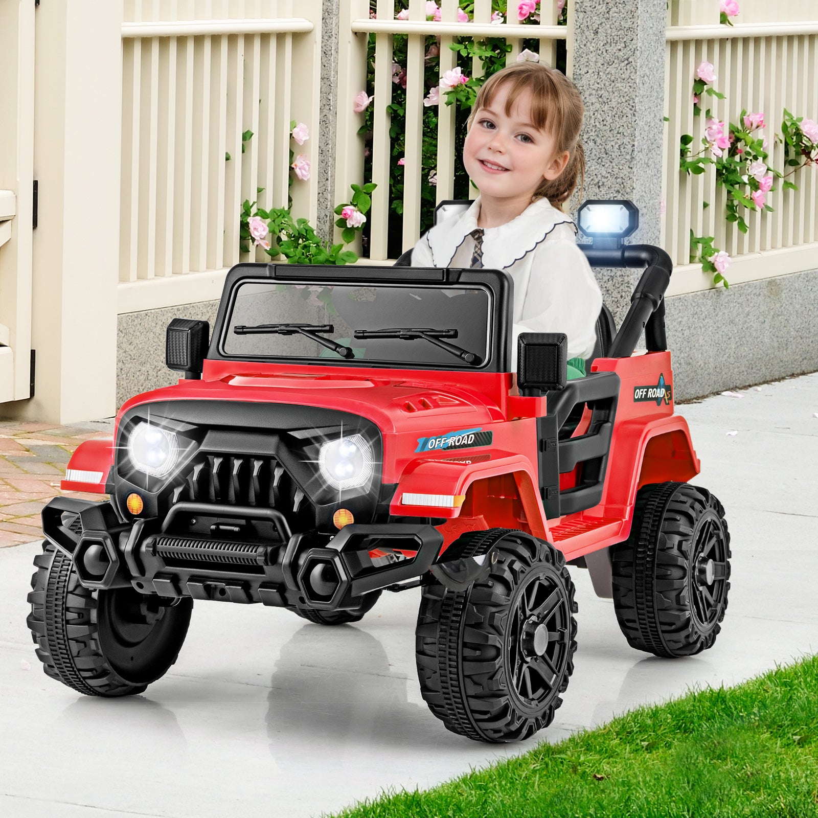 Child sitting in a red toy jeep with a white fence and flowers in the background