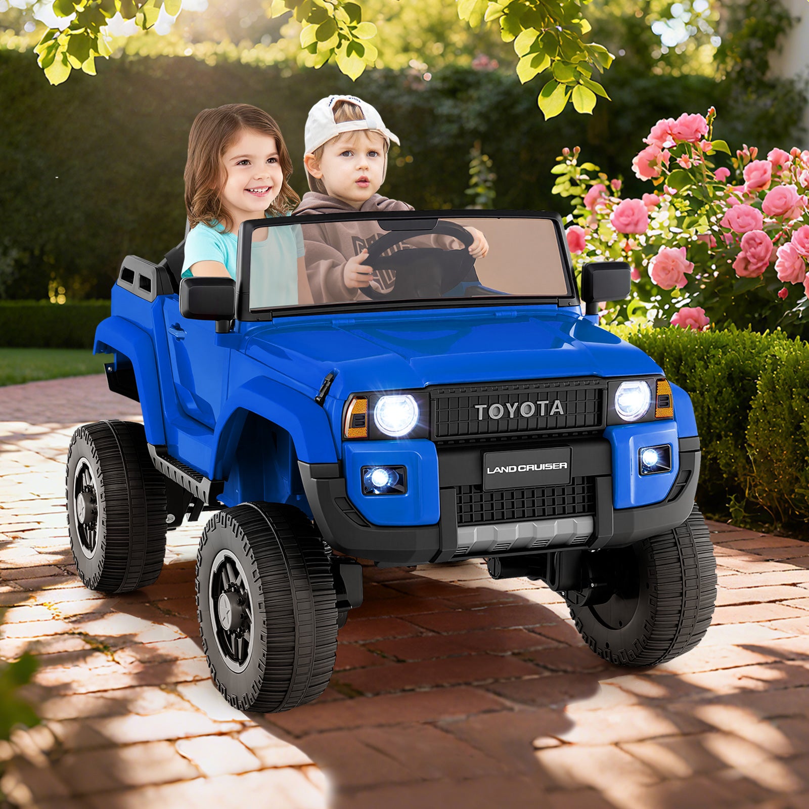 Two children in a blue toy Toyota vehicle on a brick path with flowers and greenery in the background.