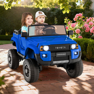 Two children in a blue toy Toyota vehicle on a brick path with flowers and greenery in the background.