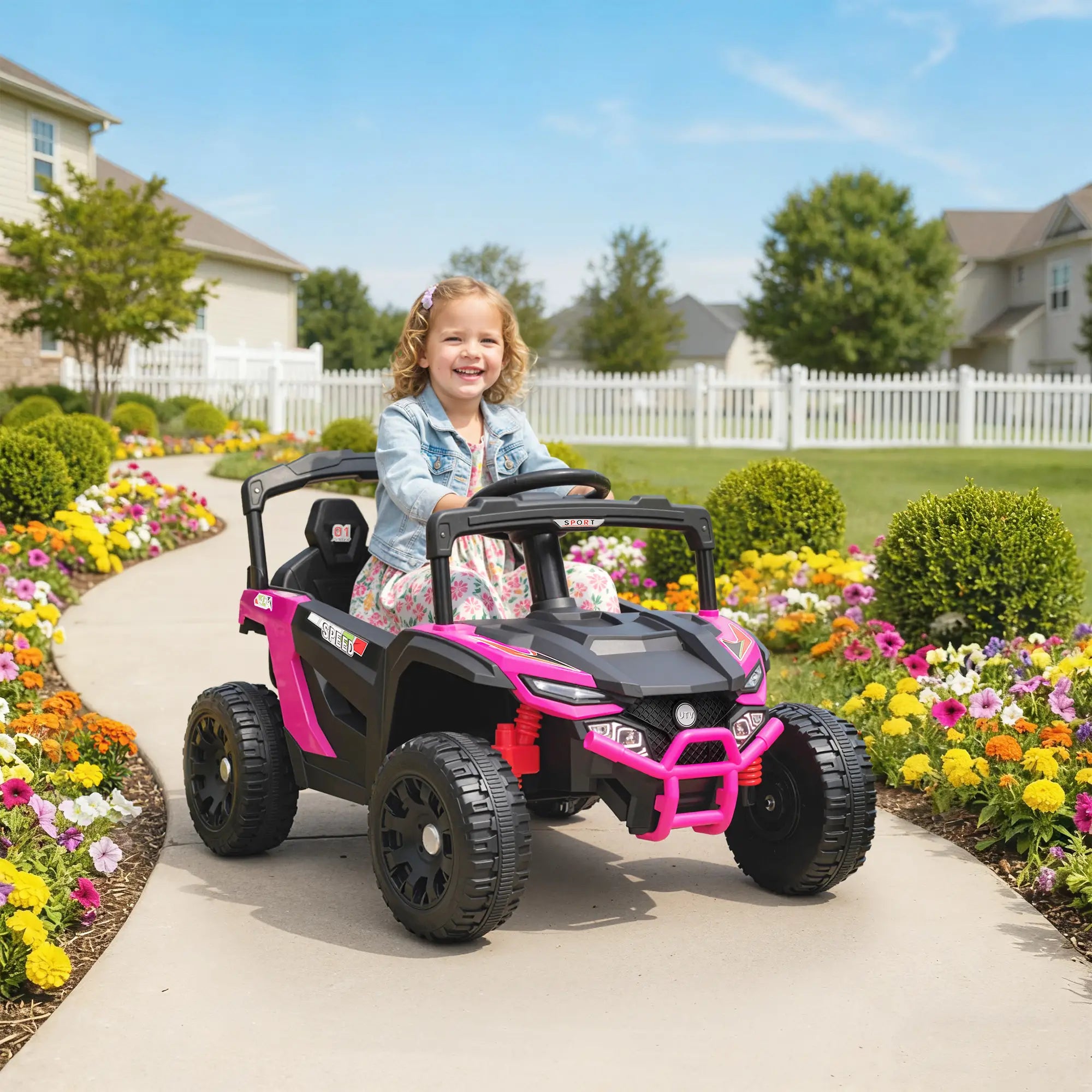 Child in a pink and black toy car on a sidewalk with a garden and houses in the background