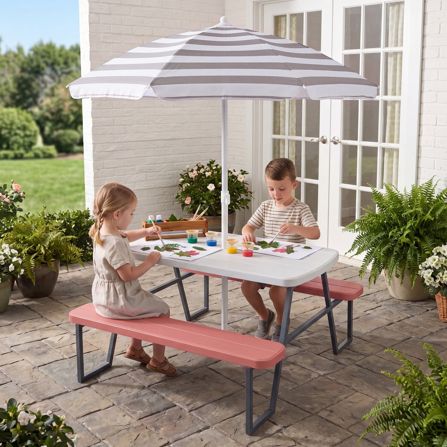 Two children sitting at a children's picnic table with a striped umbrella on a patio.
