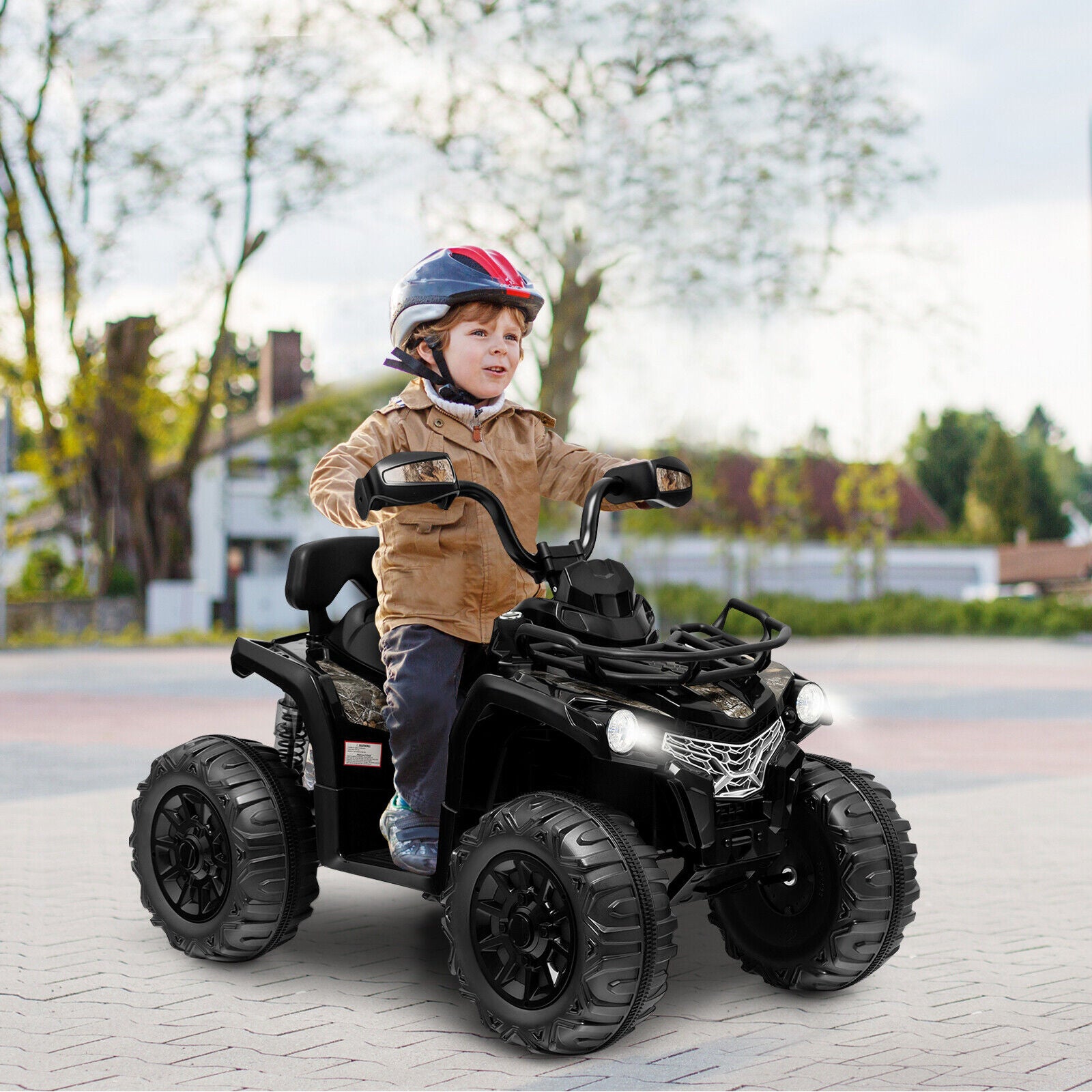 Child riding a toy ATV on a paved area with trees and houses in the background