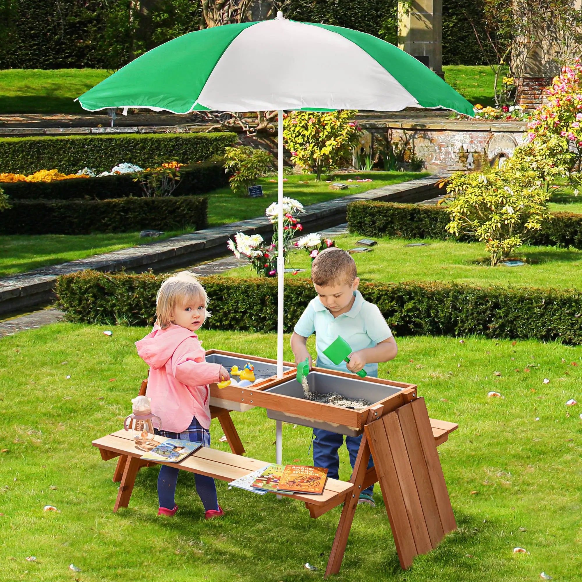 Two children playing at a sand table with a green and white umbrella in a garden setting.