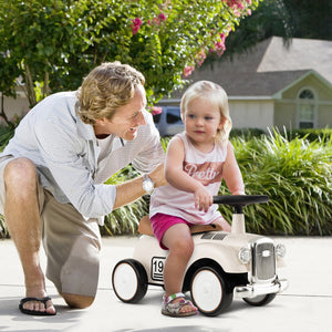 Man pushing a child in a toy car outdoors with greenery and a house in the background