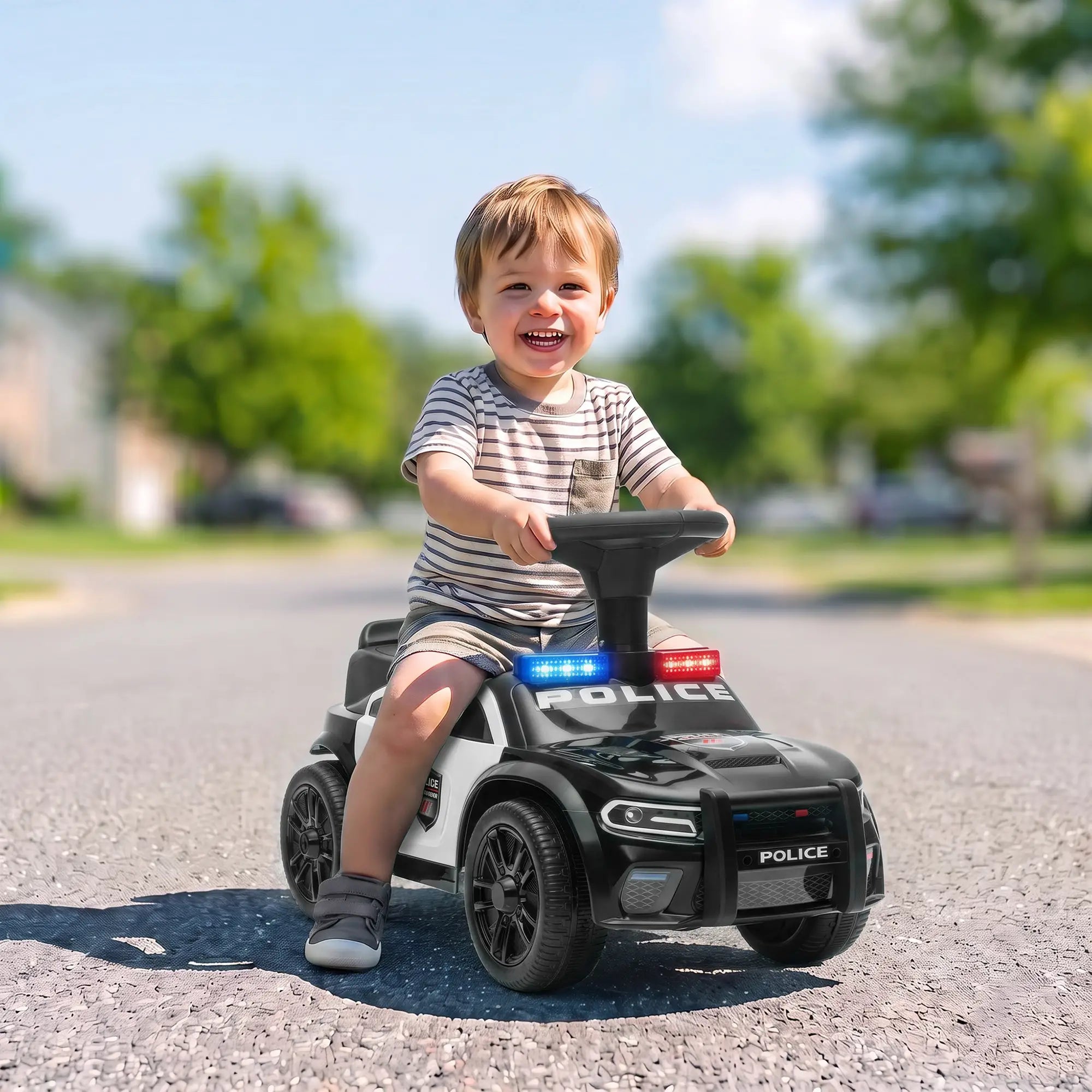 A young child riding a black and white police-themed toy car on an asphalt street outdoors. The toy car has 'POLICE' written on the hood and roof, with red and blue light accents, and the child is smiling while holding the steering wheel.