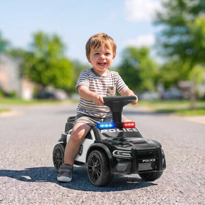 A young child riding a black and white police-themed toy car on an asphalt street outdoors. The toy car has 'POLICE' written on the hood and roof, with red and blue light accents, and the child is smiling while holding the steering wheel.