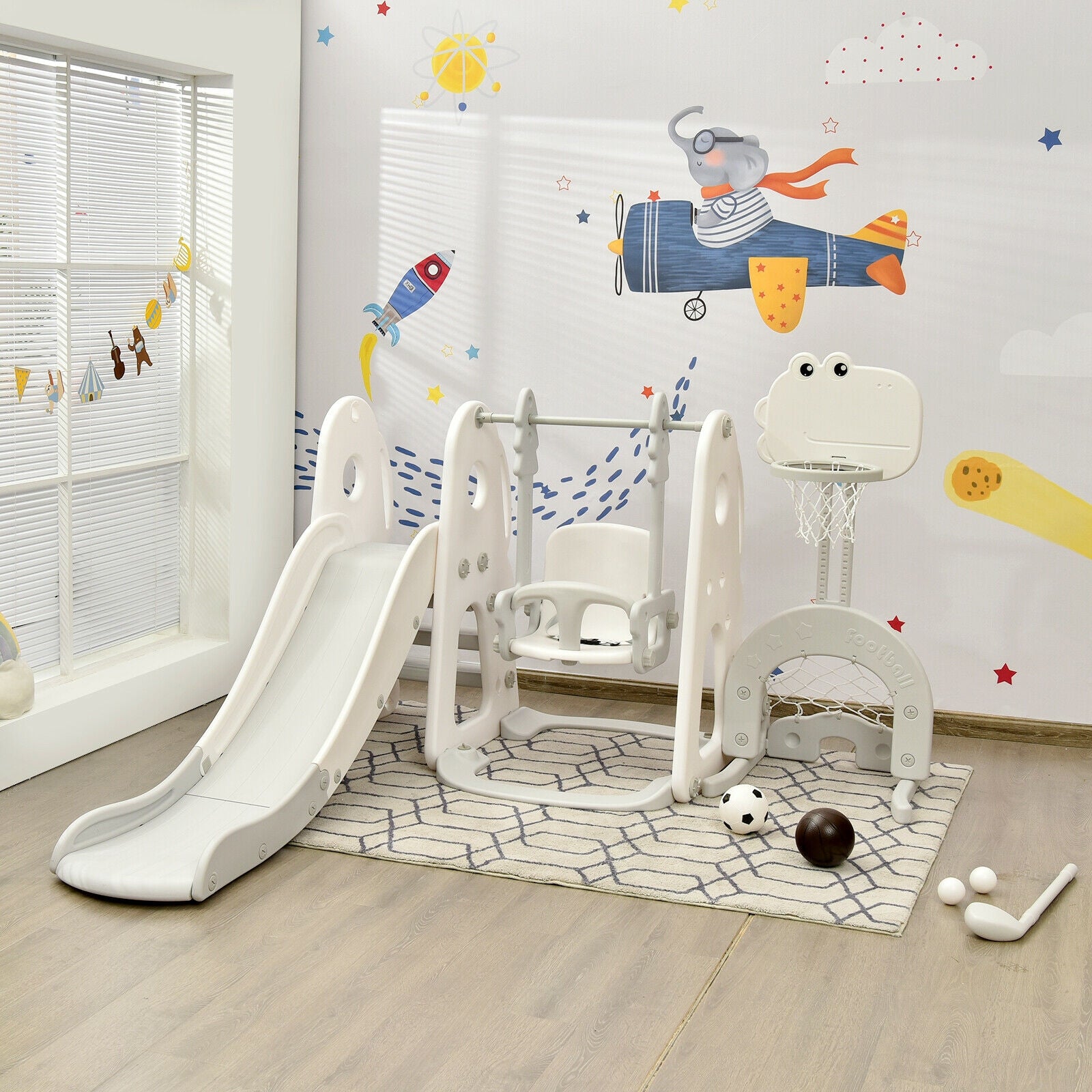 Children's indoor play area with slide, swing set, and basketball hoop against a wall with space-themed decorations.