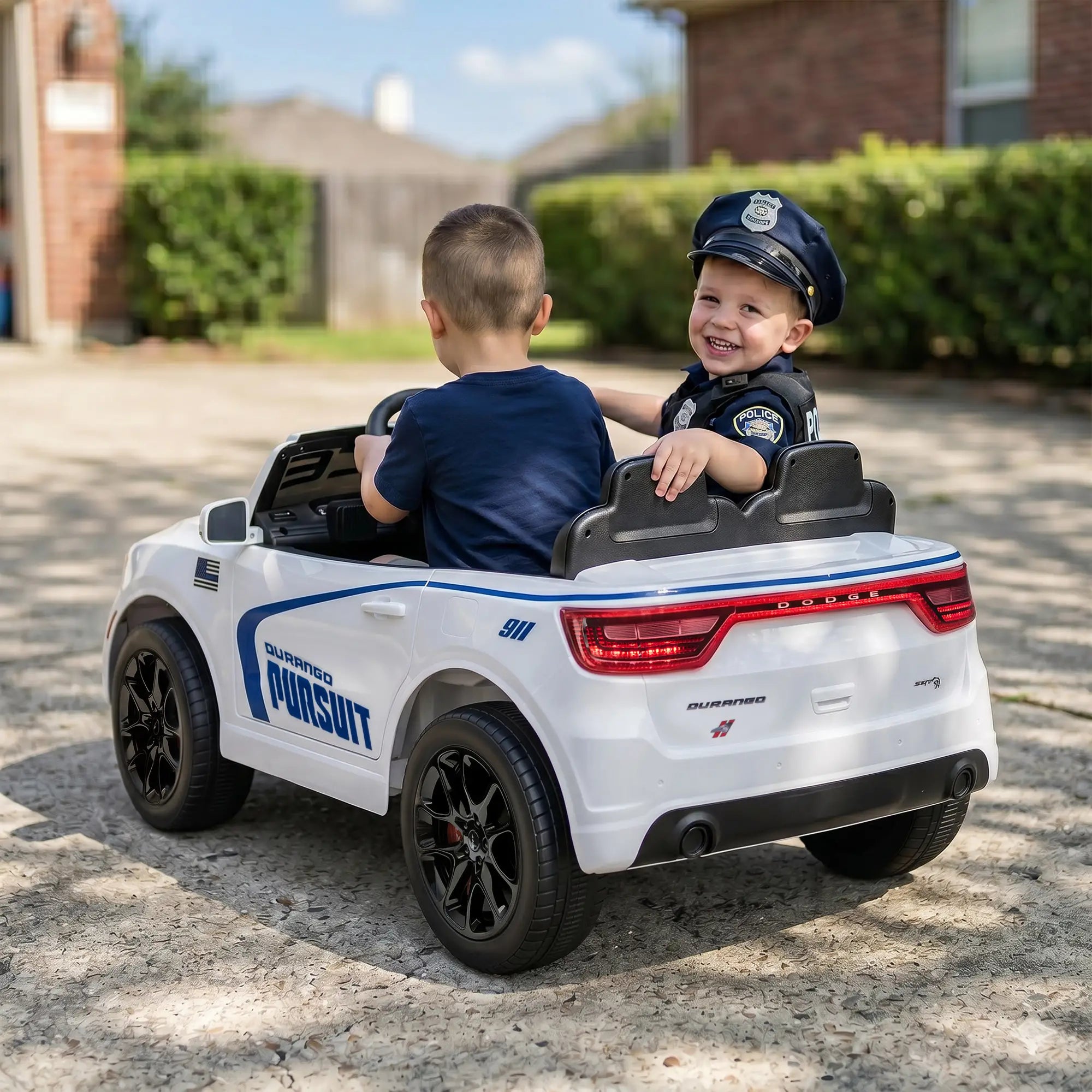 A toddler in a police hat drives a white and blue toy ride-on car outdoors on a driveway. The car has black wheels and a black seat with a smiling child sitting inside.