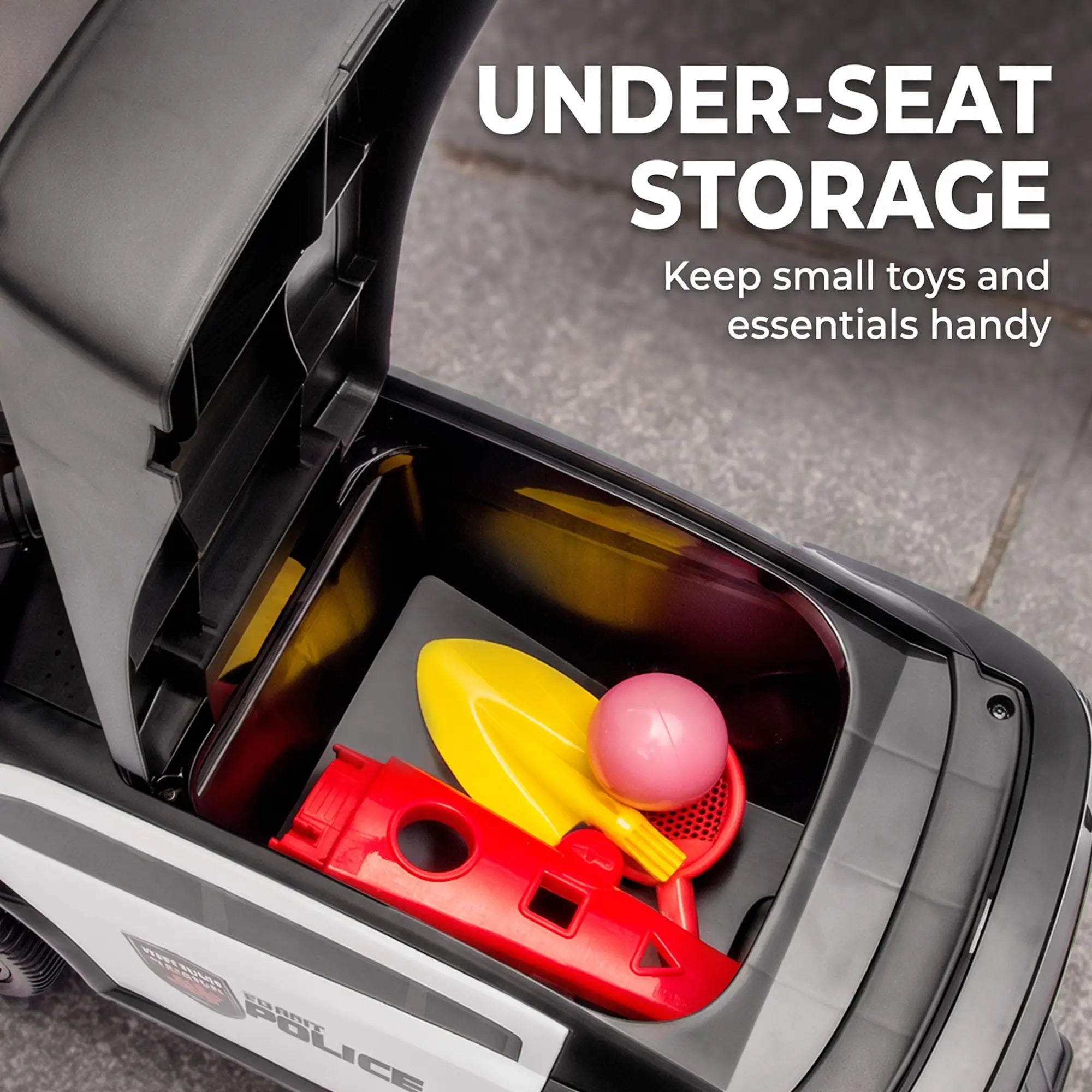 Close-up of a toy vehicle's open under-seat storage compartment containing a yellow shovel, red toy piece, and pink ball, with text reading 'UNDER-SEAT STORAGE Keep small toys and essentials handy' over a gray pavement background.