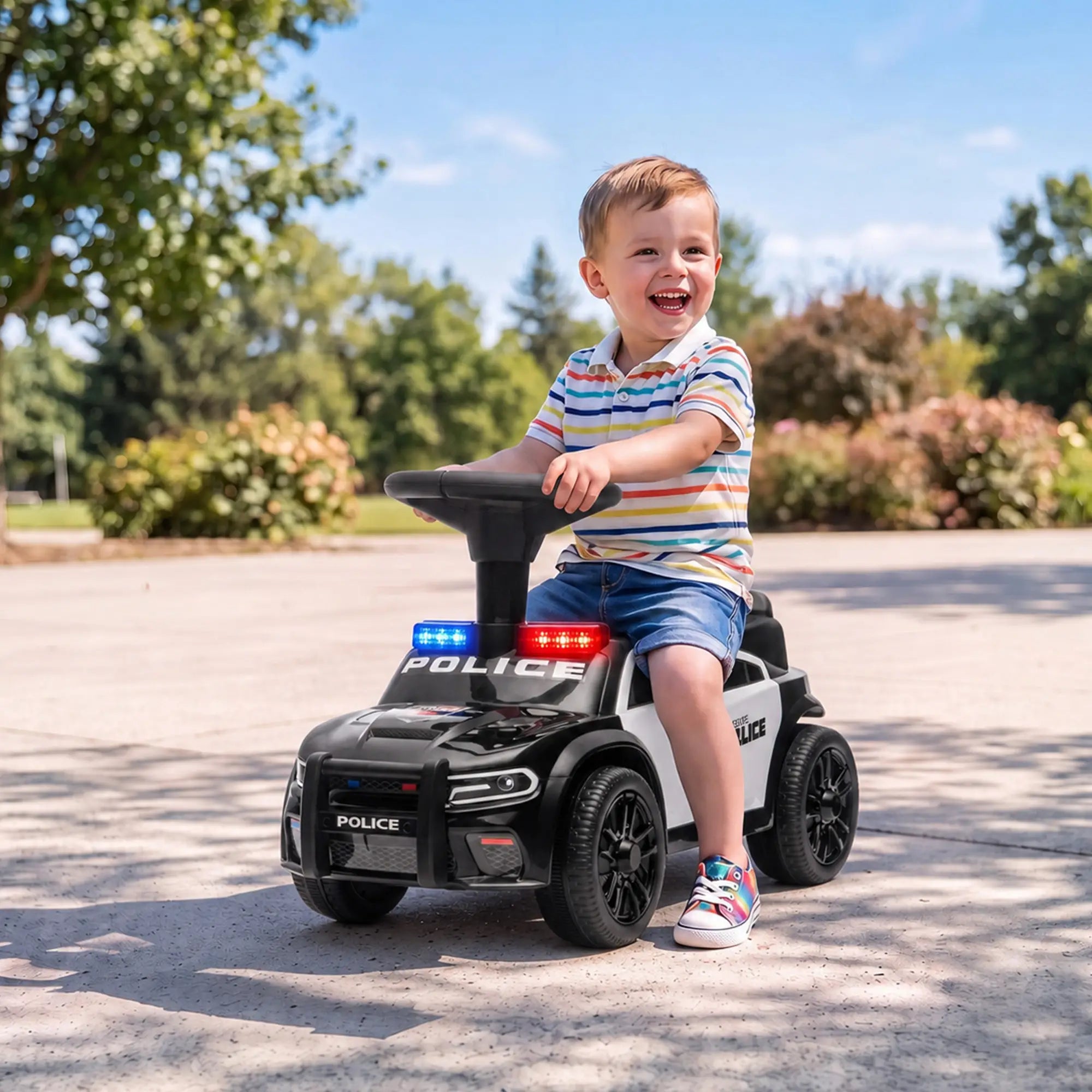 A young boy wearing a striped polo shirt and denim shorts sits smiling on a black and white police-themed ride-on toy car outdoors on a paved path.