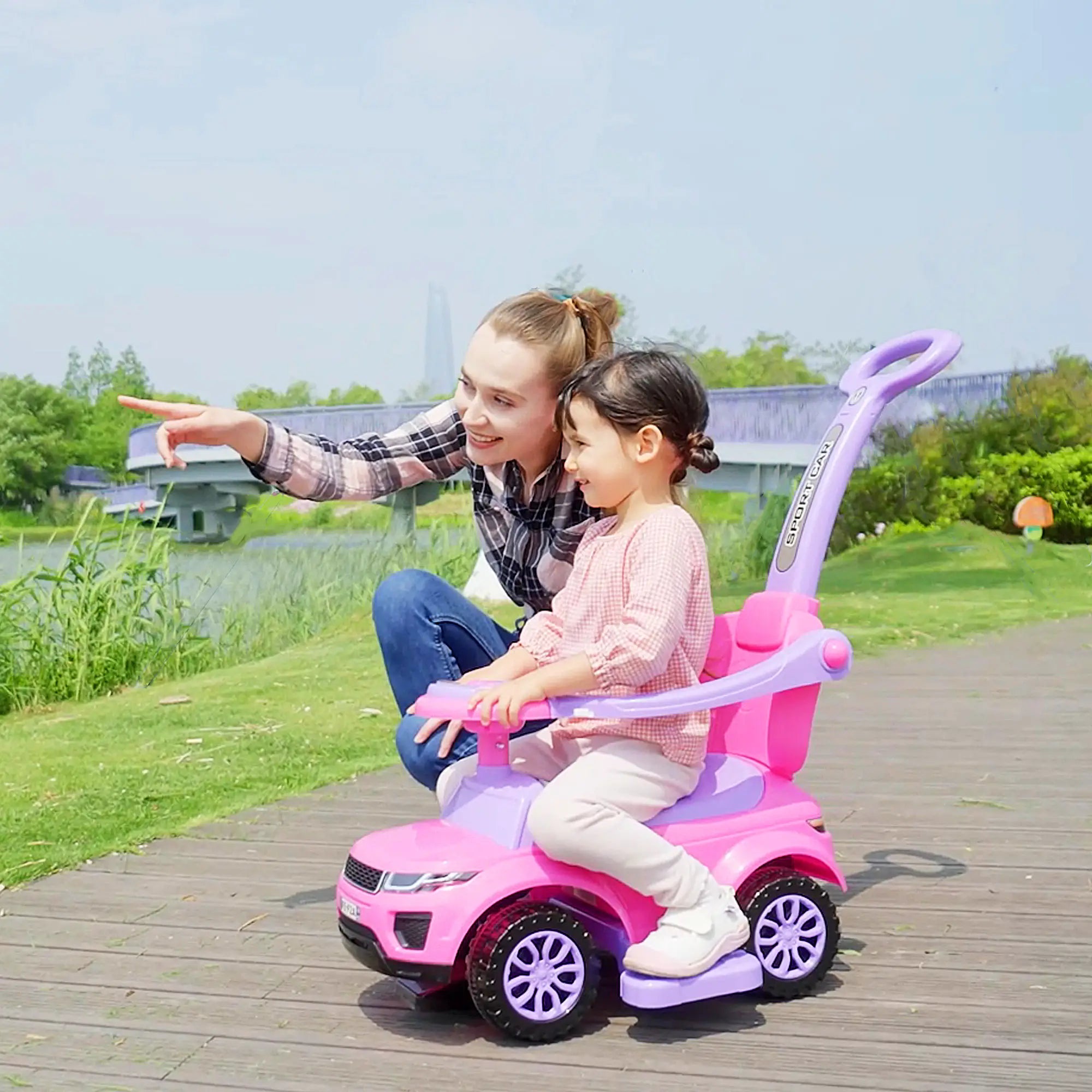 Woman and child riding a pink toy car outdoors on a wooden path.