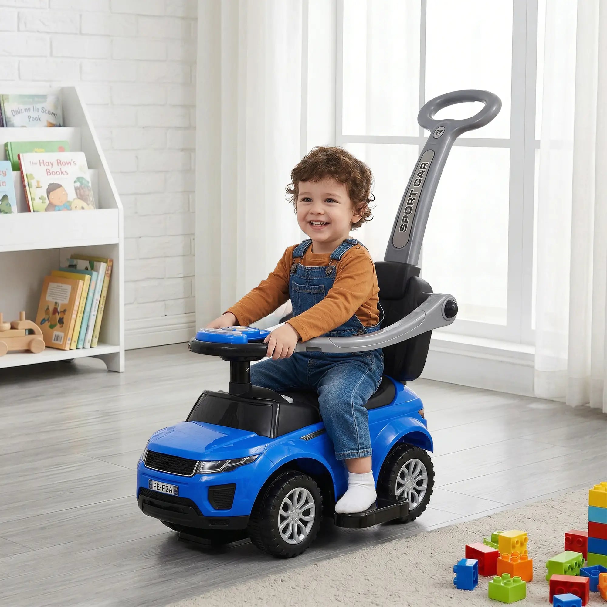 Child sitting in a blue toy car in a room with books and toys.