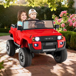 Two children in a red toy Toyota Land Cruiser vehicle on a brick path with flowers and greenery in the background.