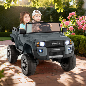Two children in a toy Toyota Land Cruiser vehicle on a brick path with flowers and greenery in the background.