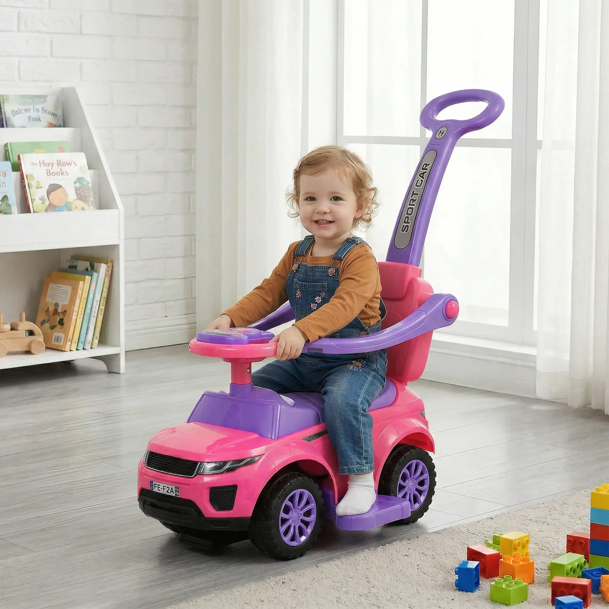 Child sitting on a pink and purple toy car in a room with books and toys.