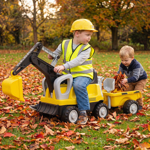 Two young children playing outdoors in a grassy park covered with fallen autumn leaves. One child sits on a yellow and black toy excavator, wearing a yellow hard hat and safety vest, while the other child pushes a small yellow toy dump truck carrying leaves.