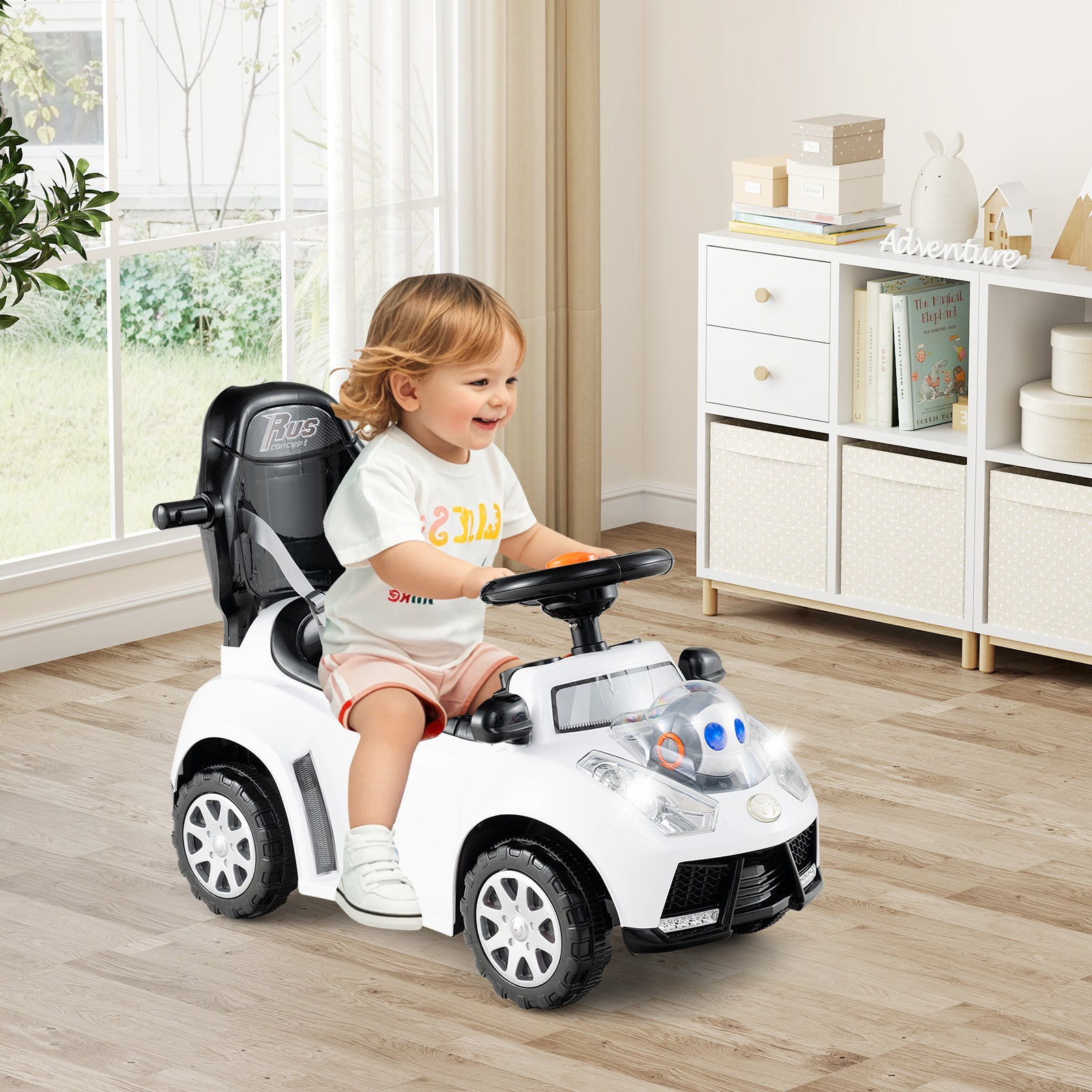 Child sitting in a toy car in a room with white furniture and large windows.