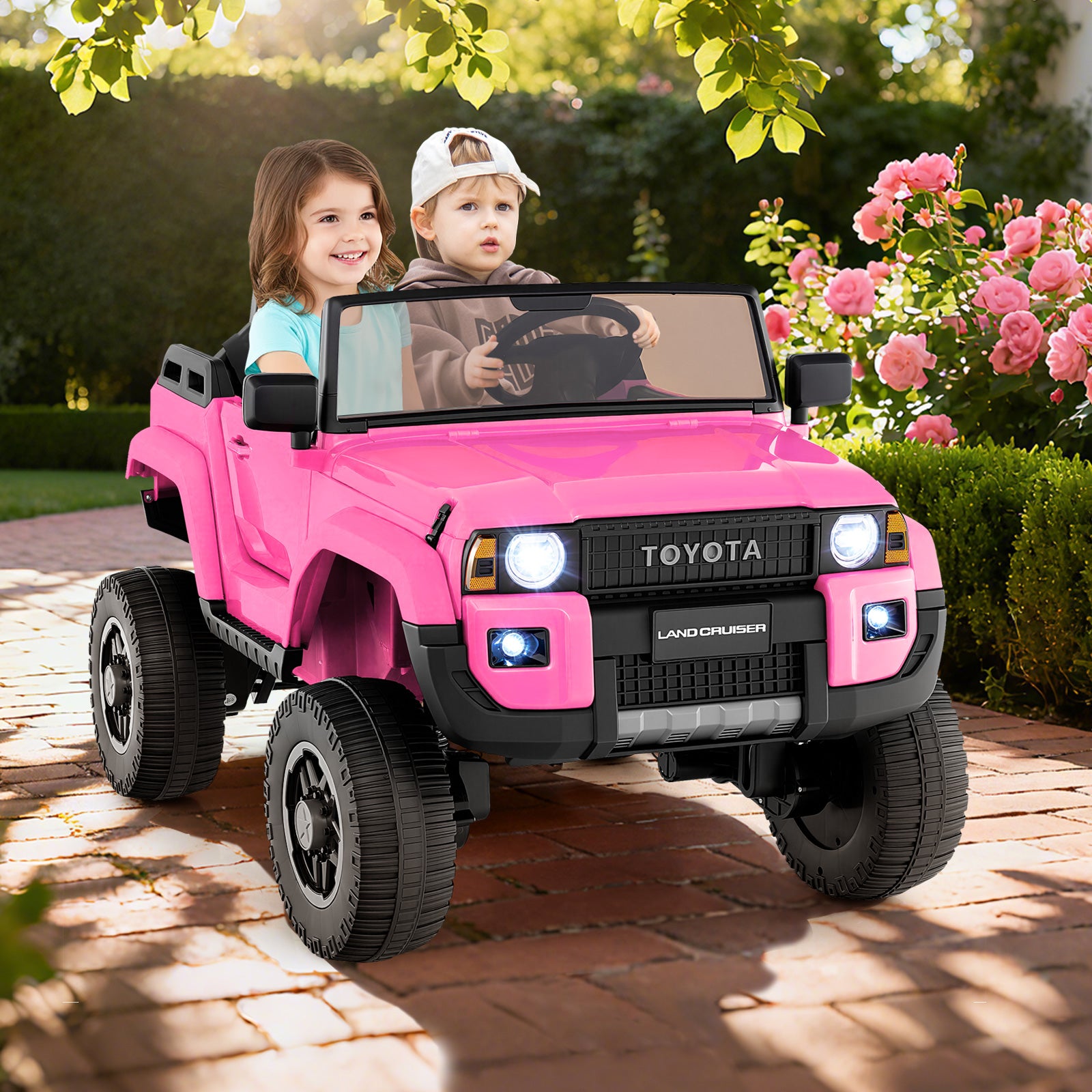 Two children in a pink toy Toyota vehicle on a brick path with flowers and greenery in the background.
