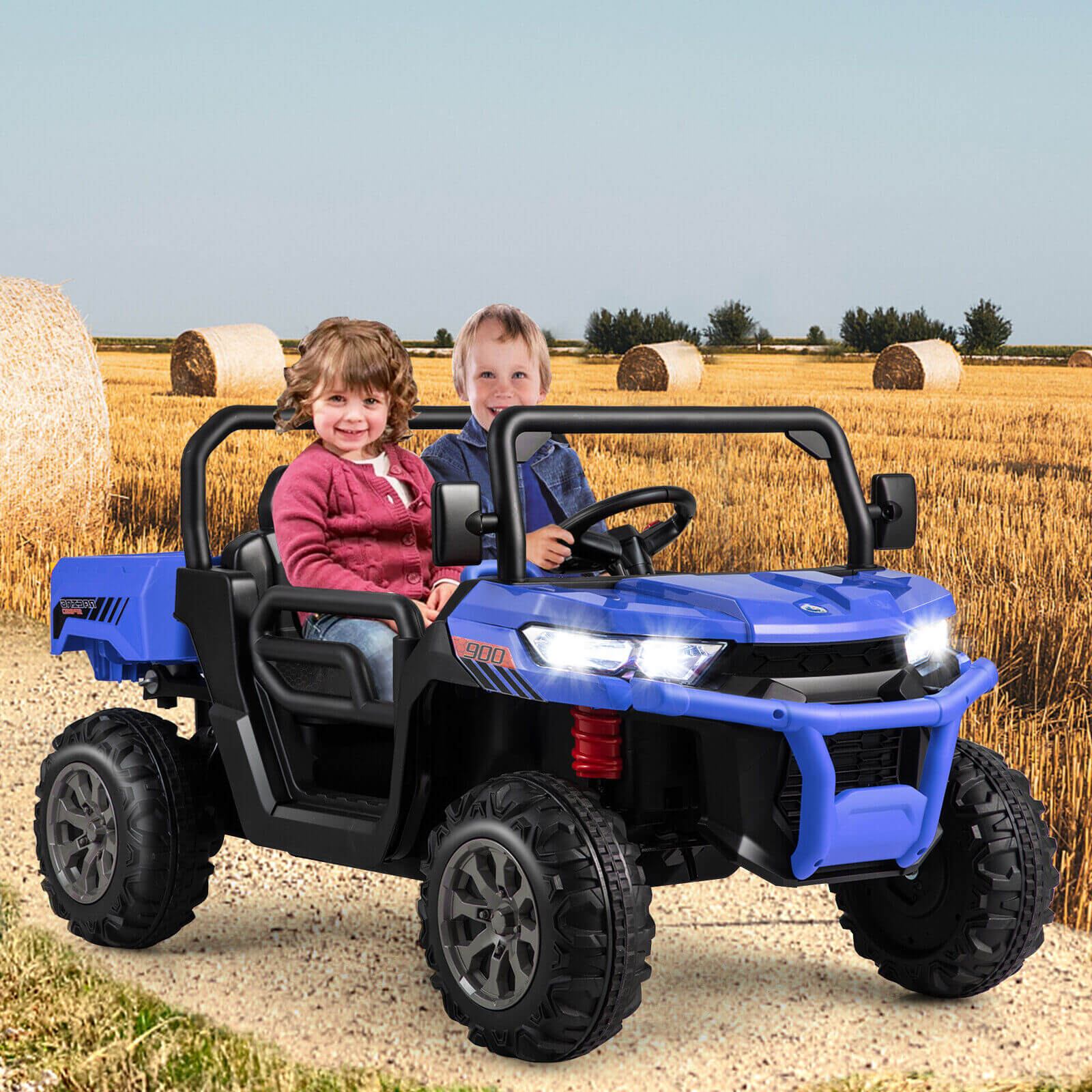 Two children sitting in a blue toy off-road vehicle in a field with hay bales.